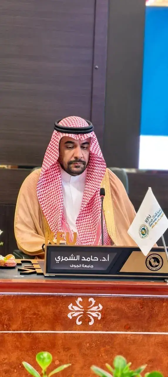 سعادة وكيل الجامعة للدراسات العليا والبحث العلمي الأستاذ الدكتور حامد مناور الشمري يجلس خلف طاولة اجتماع رسمية، أمامه ميكروفون ولافتة تعريف، وتظهر بجانبه أعلام صغيرة على الطاولة.